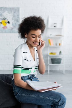 Neşeli Afro-Amerikan kadın oturma odasında akıllı telefonla konuşurken belgelere bakıyor.