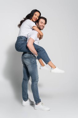 Mutlu genç adam, gri kameraya bakarken gülümseyen kız arkadaşını sırtında taşıyor.