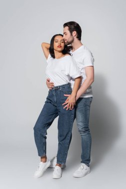 full length view of man with closed eyes hugging seductive woman in white t-shirt and jeans on grey clipart
