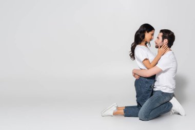 side view of couple in white t-shirts and jeans standing on knees and hugging on grey clipart