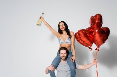 Kalp şeklinde balonları olan adam, gri üzerinde şampanya şişesi olan seksi ve mutlu kadını sırtında taşıyor.
