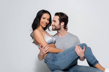 Tişörtlü mutlu adam sütyenli seksi kadını tutuyor ve gri renkli kameraya bakıyor.