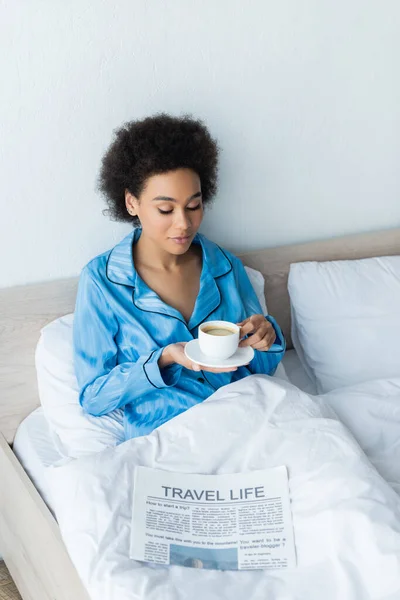 穿着睡衣的非洲裔美国妇女 在旅行生活报附近的床上拿着一杯咖啡 — 图库照片