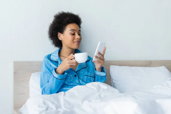 Sorrindo Mulher Americana Africana Pijama Usando Smartphone Segurando Xícara Café — Fotografia de Stock