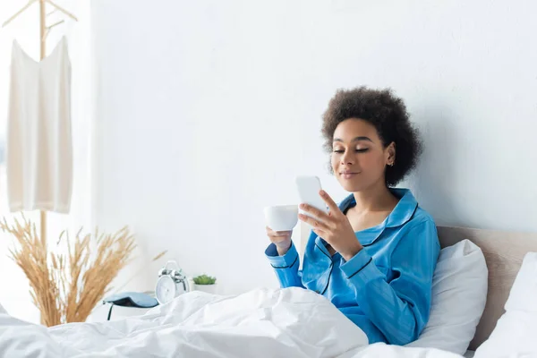 穿着睡衣 拿着手机 在床上端着咖啡的非洲裔美国女人笑着 — 图库照片