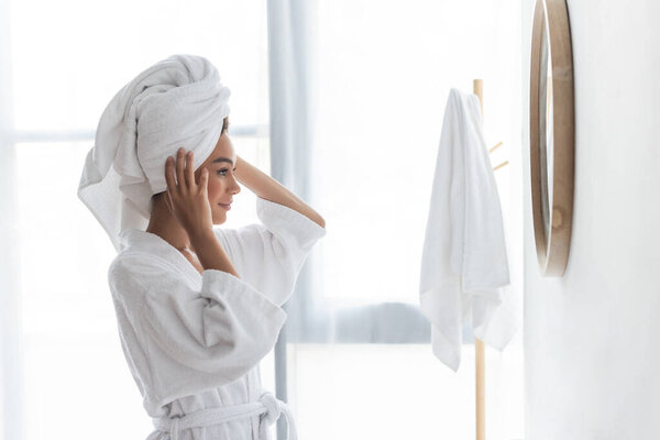 joyful african american woman adjusting towel and looking at mirror in bathroom