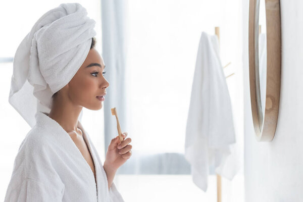 side view of african american woman looking at mirror and holding toothbrush in bathroom