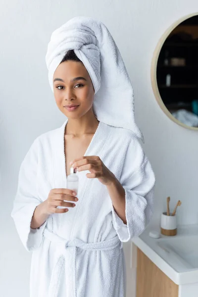 白いタオルの若いアフリカ系アメリカ人女性バスルームで血清とボトルを保持バスローブ — ストック写真