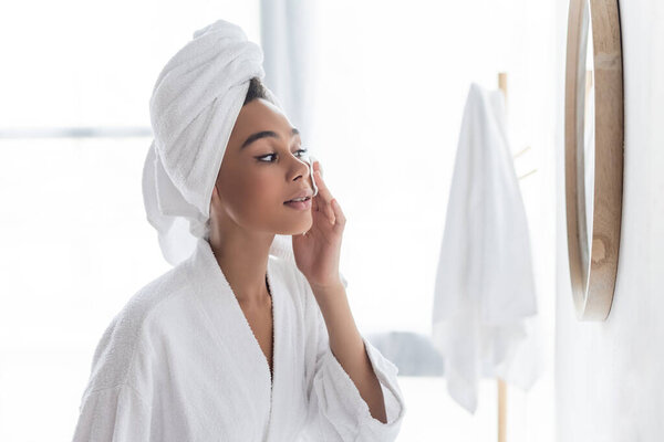 young african american woman in towel cleansing face with cotton pad near mirror