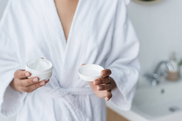 частичный вид на африканскую американку в халате, держащую контейнер с кремом для лица в ванной комнате 