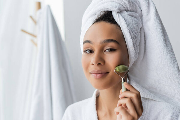 довольная африканская американка в белом полотенце массирует лицо нефритовым роллером в ванной комнате 