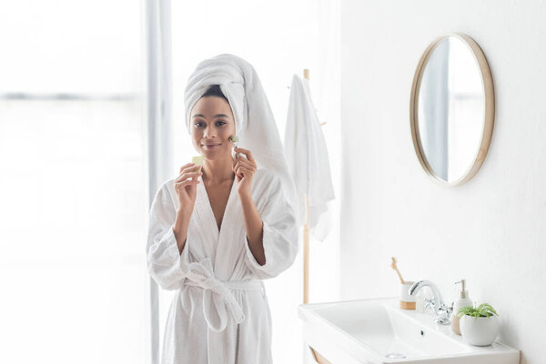 happy african american woman in bathrobe massaging face with jade roller and face scraper