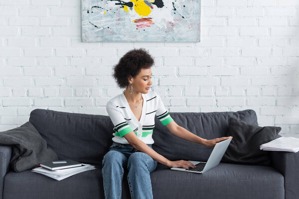 curly african american woman using laptop near papers and credit card on sofa