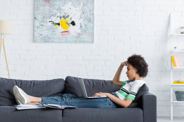 side view of curly african american woman sitting with laptop on couch