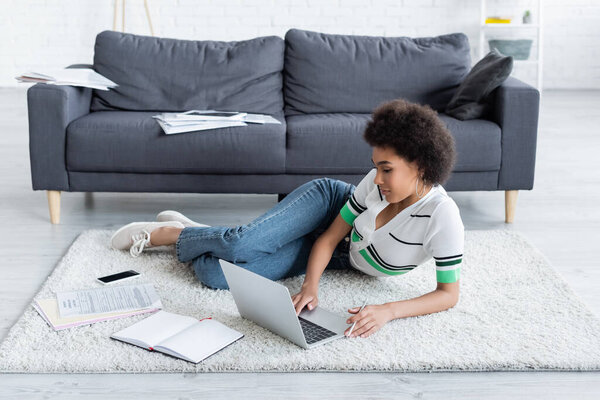 african american woman using laptop while lying on carpet 