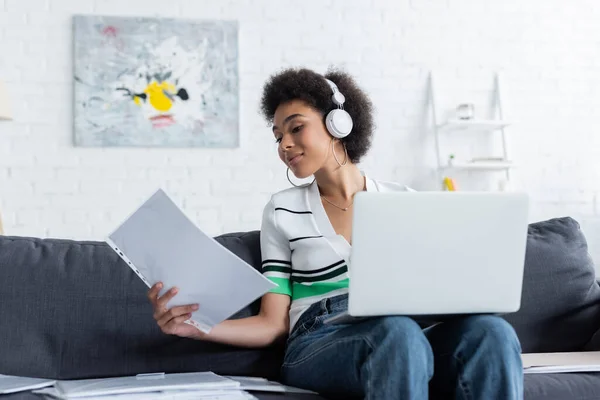 Afrykański Amerykański Freelancer Słuchawkach Bezprzewodowych Pomocą Laptopa Patrząc Papier — Zdjęcie stockowe