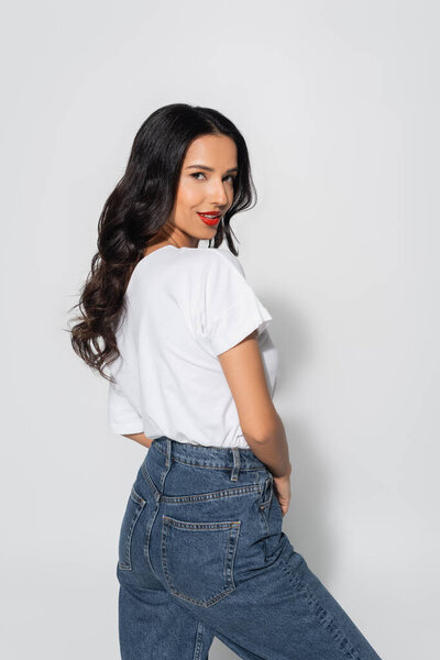 флиртующая и улыбающаяся брюнетка в белой футболке смотрит в камеру на сером