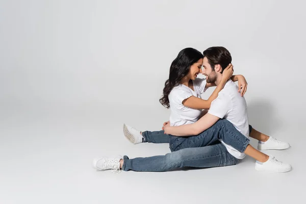 穿着白色T恤衫和牛仔裤的年轻夫妇面对面地坐在灰色的衣服上拥抱在一起 — 图库照片
