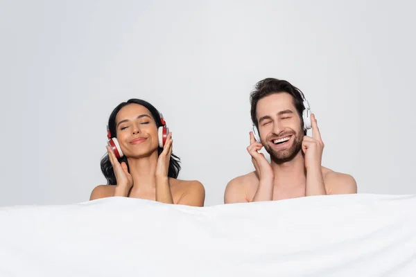 joyful couple with closed eyes listening music in headphones near white blanket isolated on grey