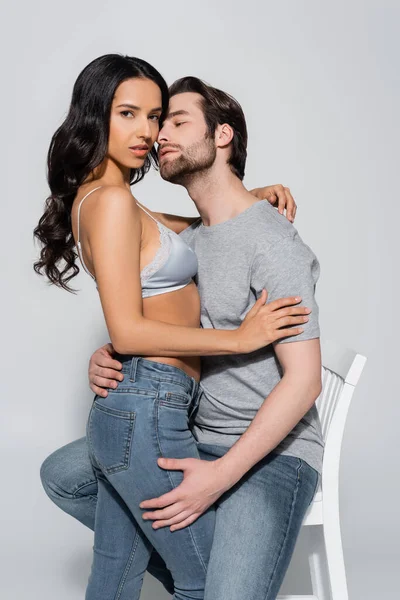 セクシー女性でジーンズとブラジャー見ますカメラで男を誘惑します上のアームチェアに座ってグレー — ストック写真