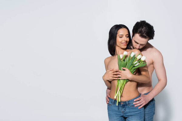 smiling woman in jeans only holding tulips near sexy shirtless man on grey