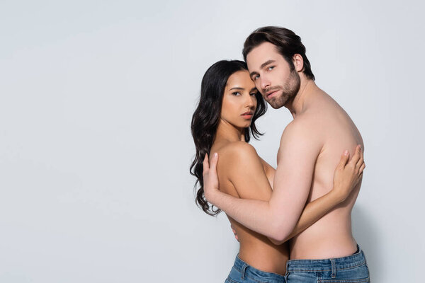 sensual couple in jeans only embracing and looking at camera on grey