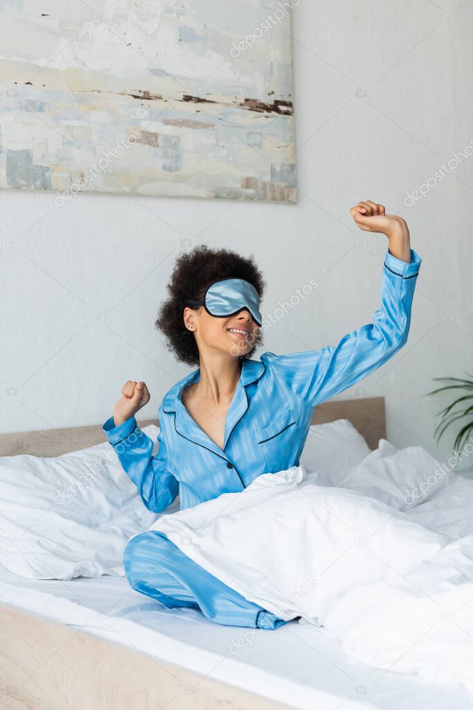 pleased african american woman in pajamas and sleeping mask stretching on bed