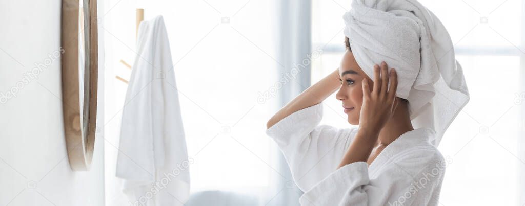joyful african american woman adjusting towel and looking at mirror in bathroom, banner