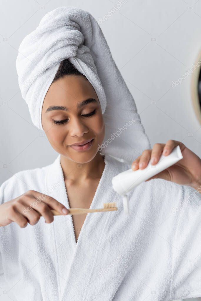 joyful african american woman in bathrobe and towel holding toothpaste and toothbrush in bathroom 