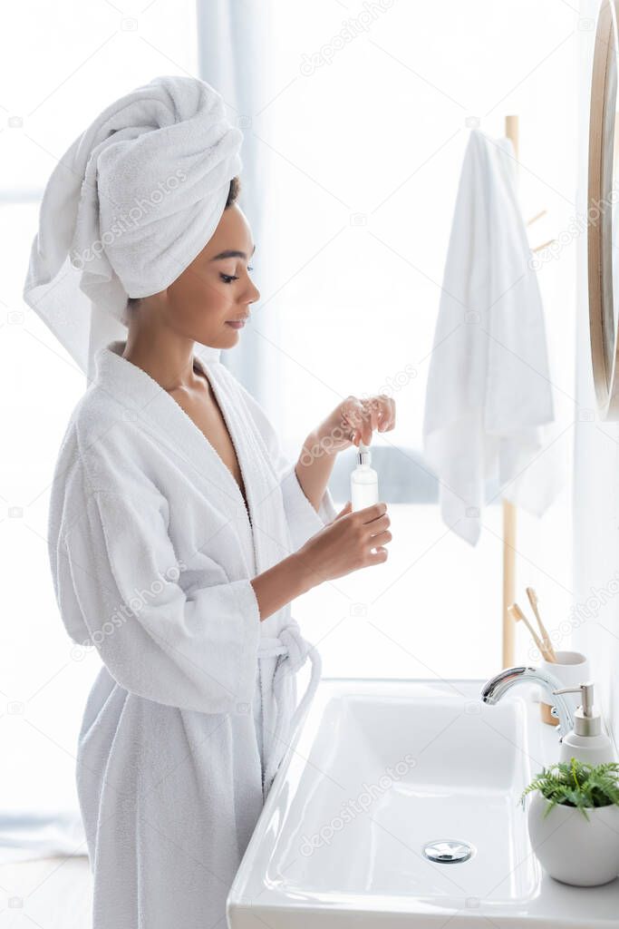 side view of young african american woman in white bathrobe holding bottle with serum in bathroom 