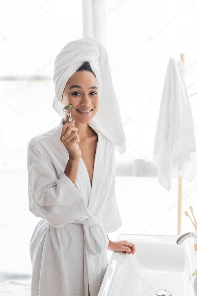 happy african american woman in bathrobe and towel massaging face with jade roller in bathroom 