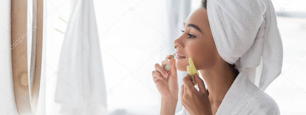 side view of african american woman in bathrobe massaging face with jade roller and face scraper, banner