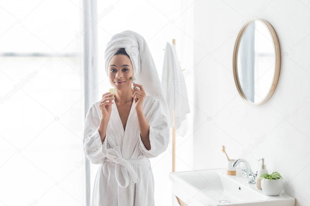 happy african american woman in bathrobe massaging face with jade roller and face scraper