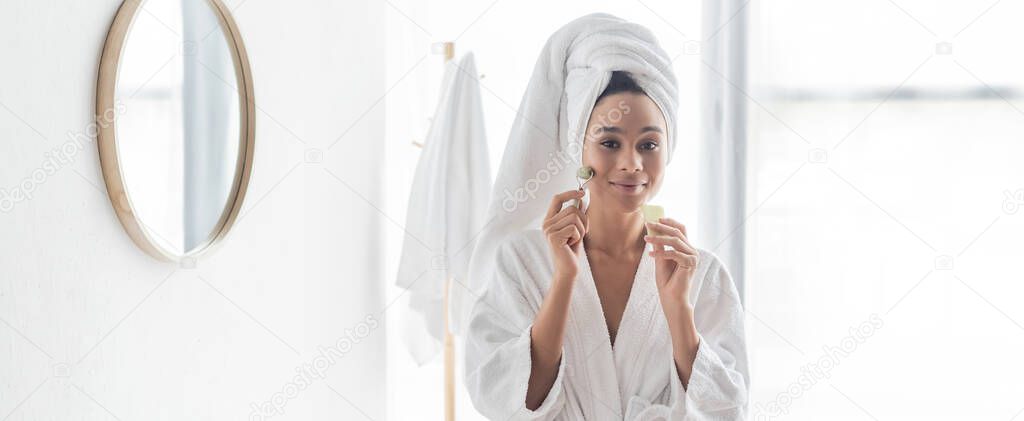 happy african american woman in bathrobe massaging face with jade roller and face scraper, banner