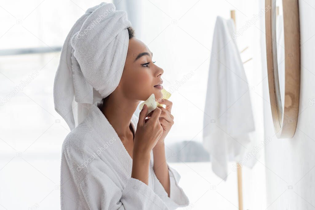 side view of young african american woman in white towel scrapping face with jade face scrapers and looking at mirror 