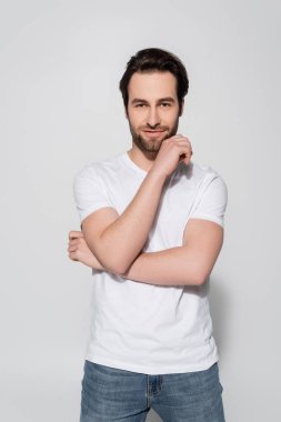 Beyaz tişörtlü pozitif bir adam. Gri renkli kameraya bakarken yüzünün yanında el ele tutuşuyor.