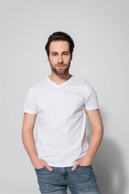 Beyaz tişörtlü esmer adam elleri gri kot pantolon cebinde duruyor.