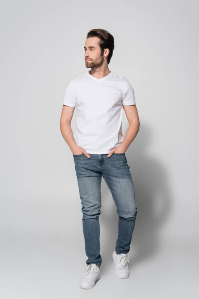 full length view of young man in white t-shirt looking away while standing with hands in pockets of jeans on grey