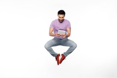 Kotlu ve mor tişörtlü mutlu adam beyaz üzerinde dijital tablet kullanırken havada süzülüyor. 
