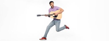 Beyaz pankartta akustik gitar çalarken havada süzülen kot pantolonlu ve mor tişörtlü bir adam.