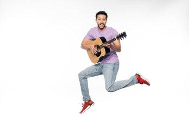 Mavi kotlu ve mor tişörtlü adam beyaz üzerinde akustik gitar çalarken havada süzülüyor. 