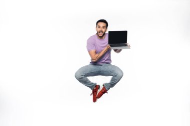 Kotlu ve mor tişörtlü pozitif adam dizüstü bilgisayarı beyaz ekranla tutarken havada duruyor. 