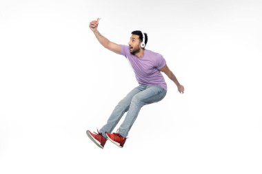 Kablosuz kulaklıklı neşeli adam selfie çekiyor ve beyaz ışıkta uçuyor.