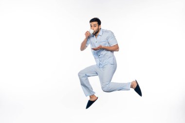 Pijama giymiş sakallı bir adam elinde fincanla beyaz havada süzülürken kahve içiyor. 