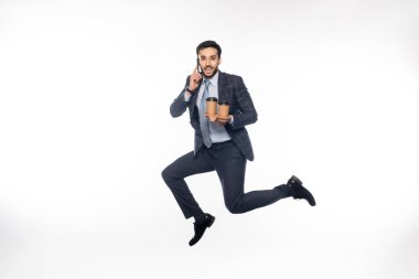 Takım elbiseli neşeli bir işadamı elinde kağıt bardaklıkla zıplıyor ve beyaz bardakla akıllı telefondan konuşuyor. 