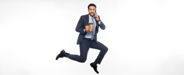 Takım elbiseli neşeli bir işadamı elinde kağıt bardaklıkla zıplıyor ve akıllı telefonuyla beyaz pankartta konuşuyor.