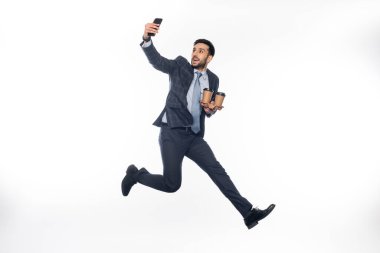 Takım elbiseli neşeli bir işadamı elinde kağıt bardakla zıplıyor ve beyaz bardaktan selfie çekiyor.