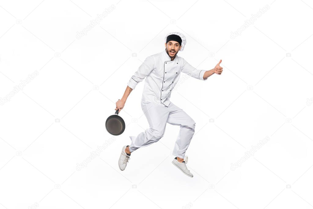 cheerful chef in hat and uniform jumping, showing thumb up while holding frying pan isolated on white 