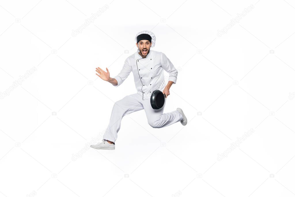 amazed chef in hat and uniform jumping and holding frying pan isolated on white 