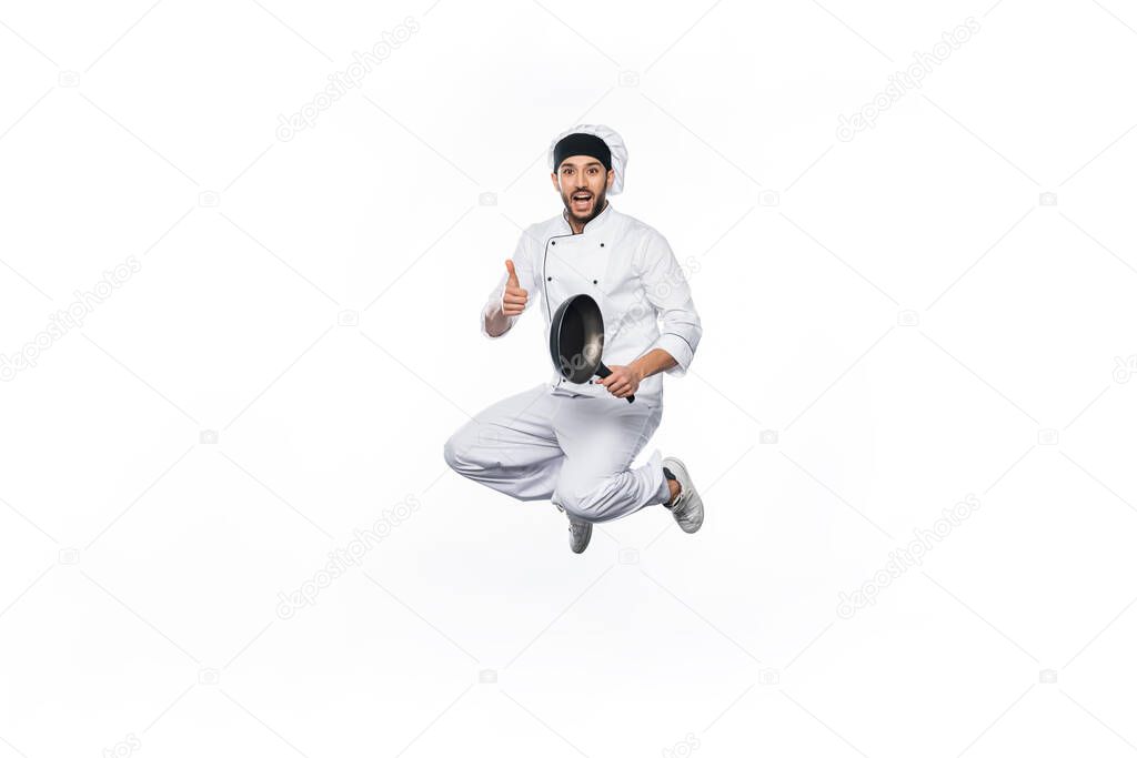 joyful chef in hat and uniform jumping, showing thumb up while holding frying pan isolated on white 